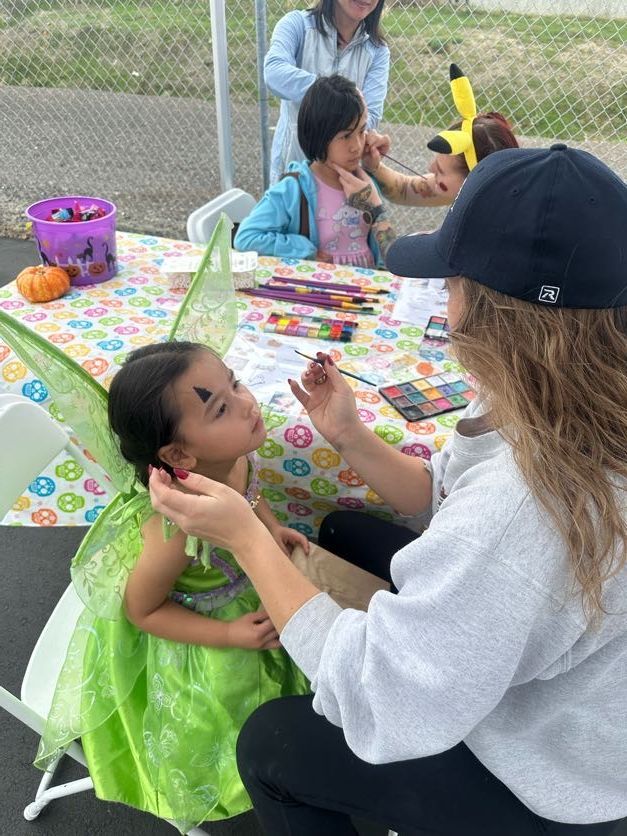 Person paints a fairy's face at an outdoor event, other children wait. Table with art supplies, pumpkin, decorations.