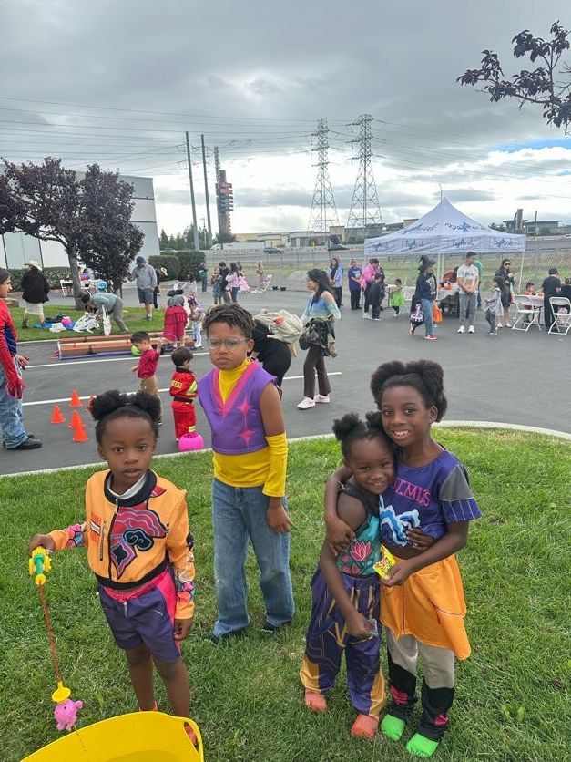 Children in colorful costumes at an outdoor event, some holding toys. Cloudy sky, power lines.