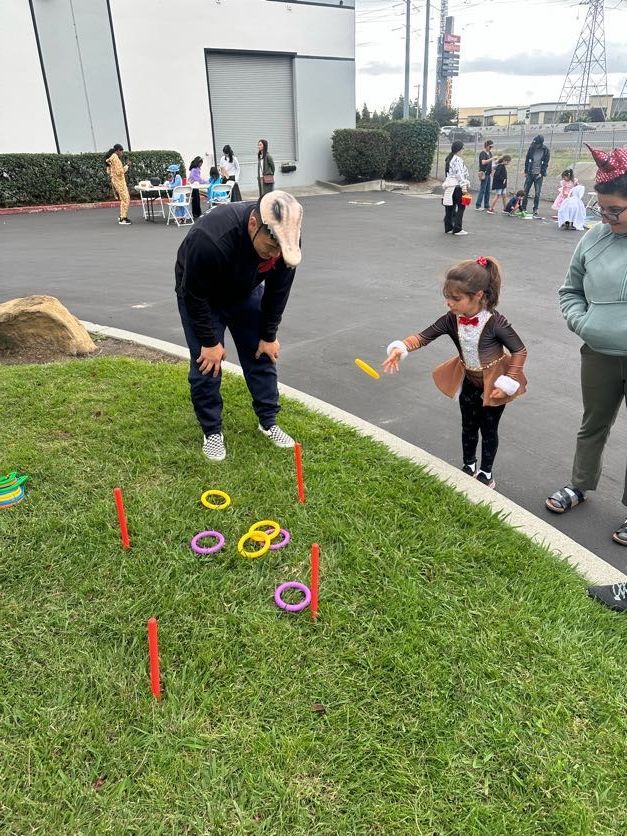 Child in costume throws ring at pegs in outdoor game, supervised by an adult.