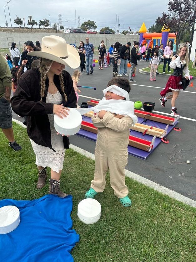 Person guiding a blindfolded child at an outdoor game with obstacles and prizes.