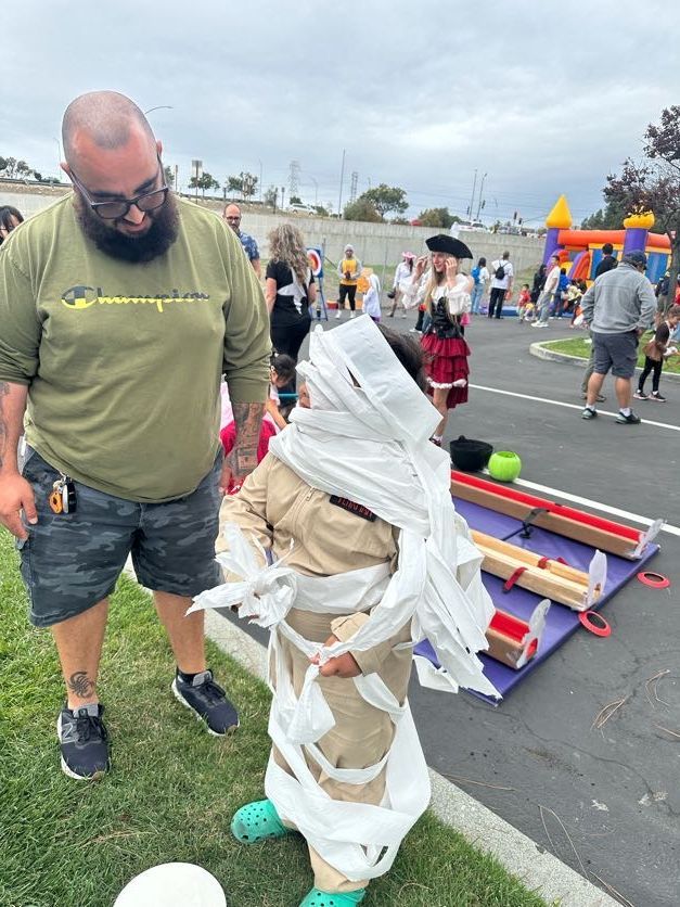 Man watches child wrapped in toilet paper at outdoor event.