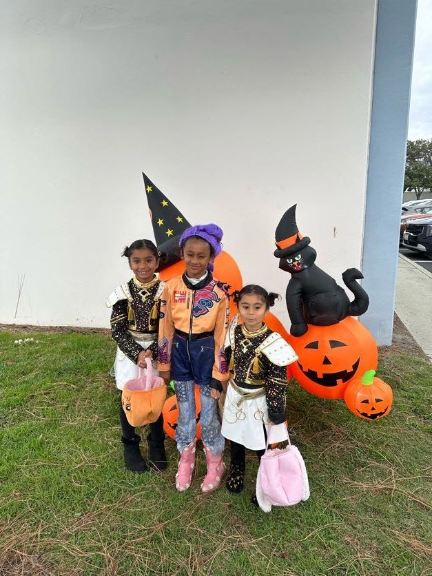 Three children in costumes pose with Halloween decorations, smiling.