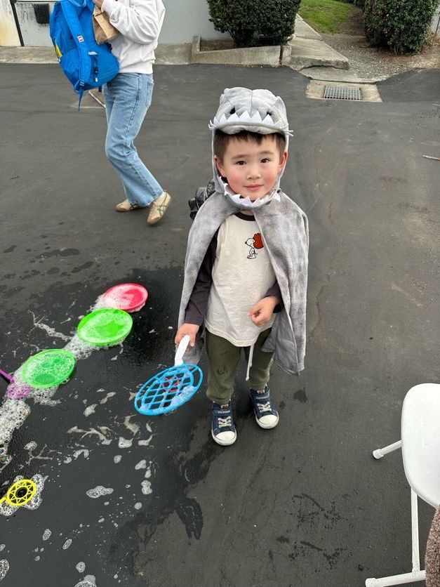 Child in a dinosaur costume holds a bubble wand, standing on a dark surface near bubbles; another person walks past.