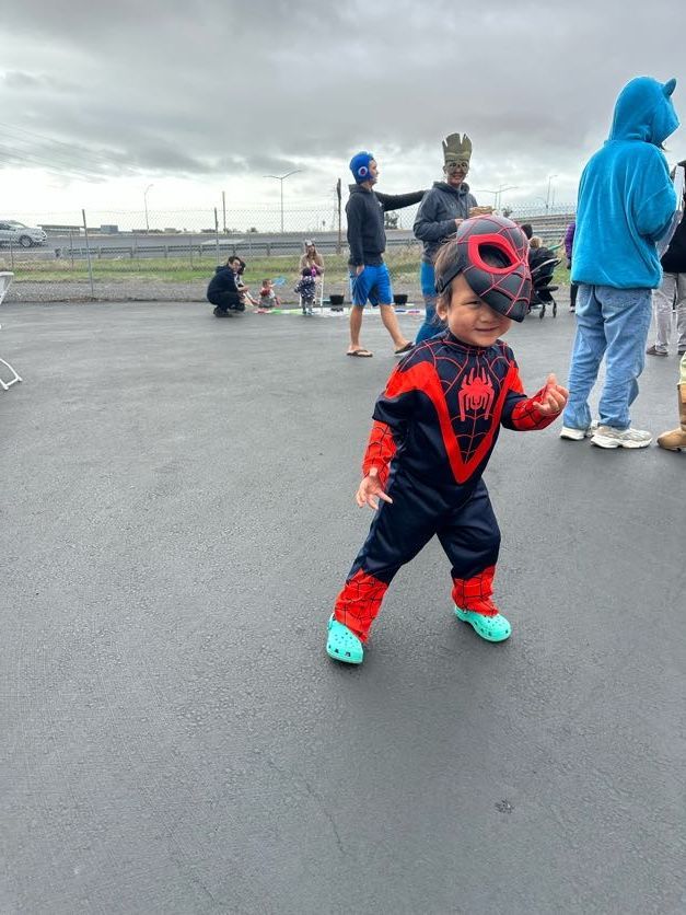 Child in Spider-Man costume, red and black, smiles. Outdoors, wet pavement, other costumed people and adults visible.