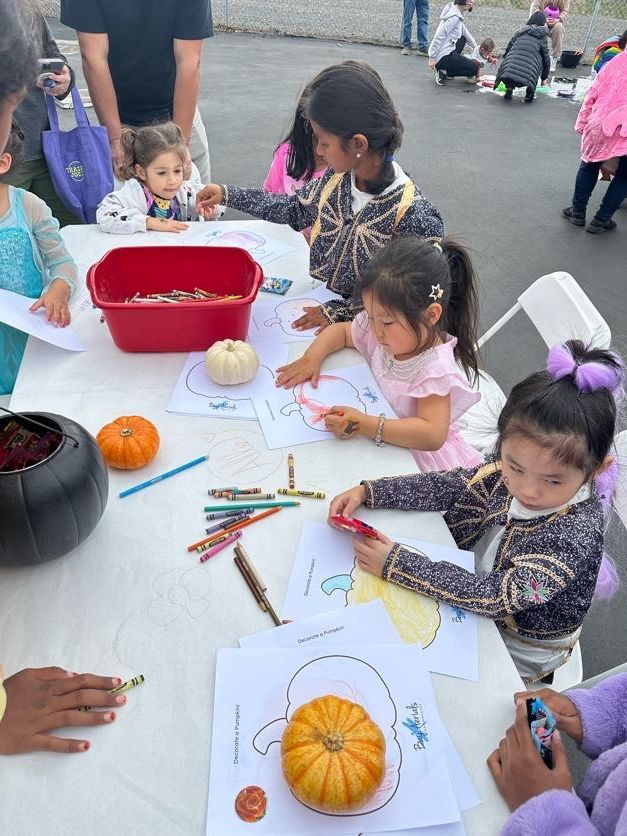 Children coloring pumpkin outlines at a table with crayons and small pumpkins. Outdoor setting.