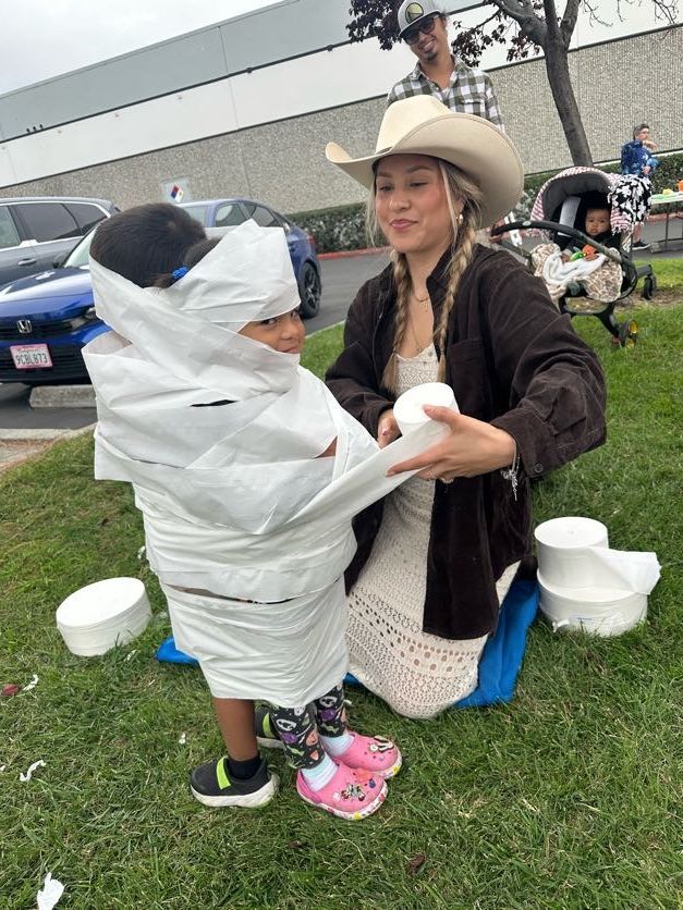 Woman wrapping child in toilet paper outdoors. Another person and a baby in a stroller are visible.