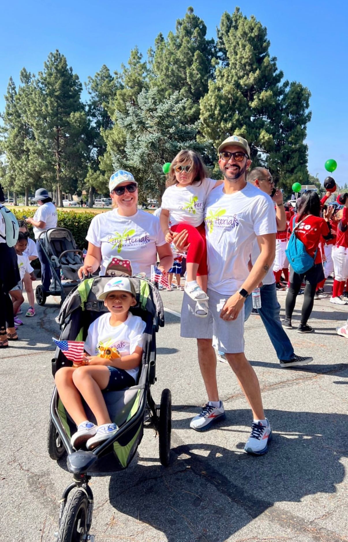Family poses outdoors: parents and two children, one in a stroller, smiling in the sunlight.