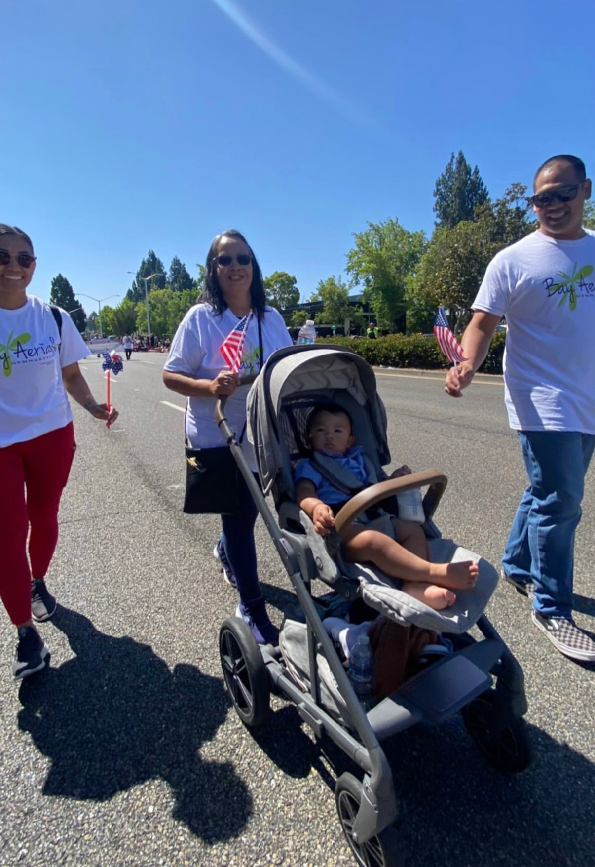 People in white shirts and holding flags walk down a sunny street, one pushing a stroller with a child.