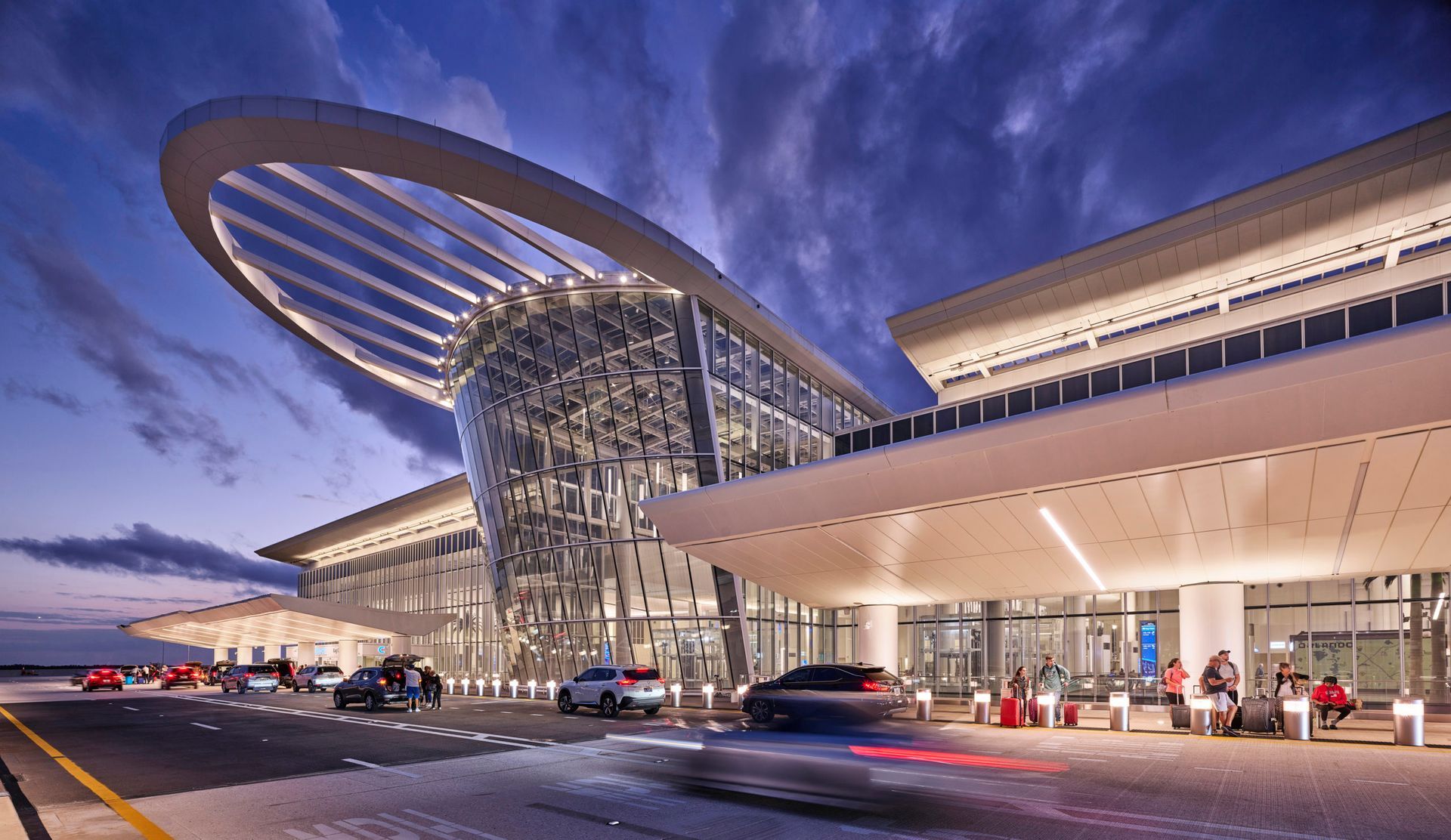 Modern airport terminal at dusk with a curved roof, glass facade, and cars at the curbside drop-off.
