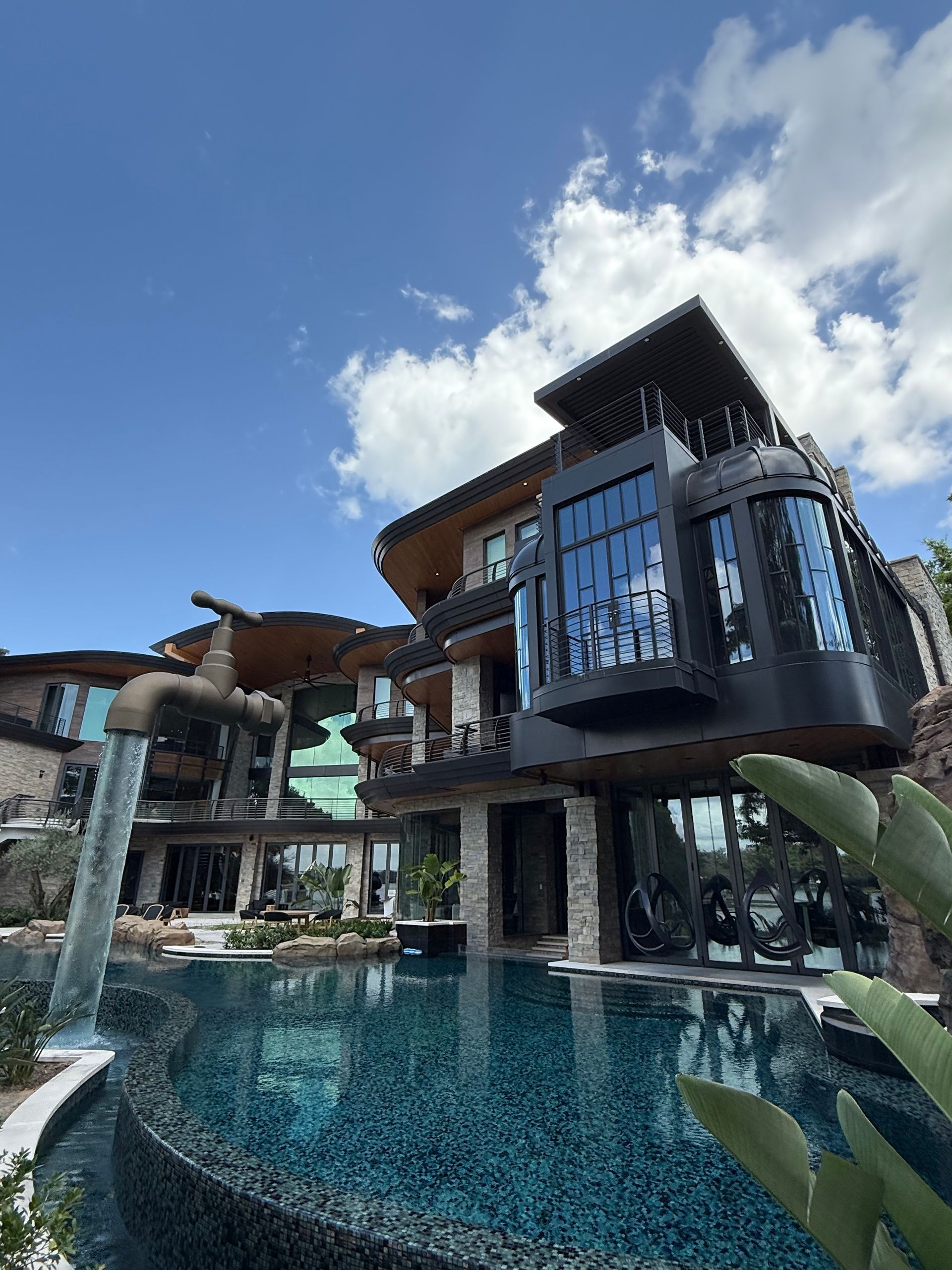 A luxurious home with a unique floating tap fountain pouring water into a swimming pool under a bright, sunny sky.