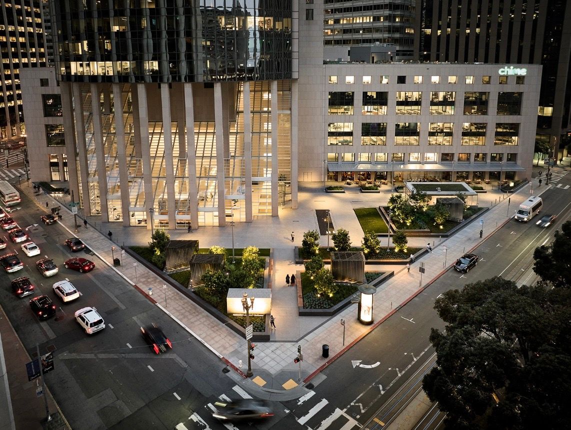 High-angle view of a modern building with a lit plaza and greenery at a street intersection at dusk, with traffic nearby.