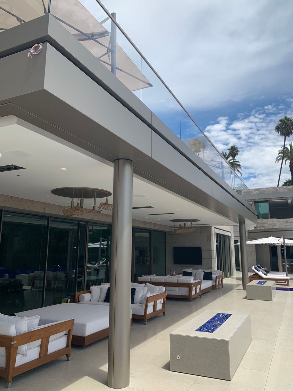 Open-air modern patio with white sofas, wooden tables, and a poolside view under a cloudy sky