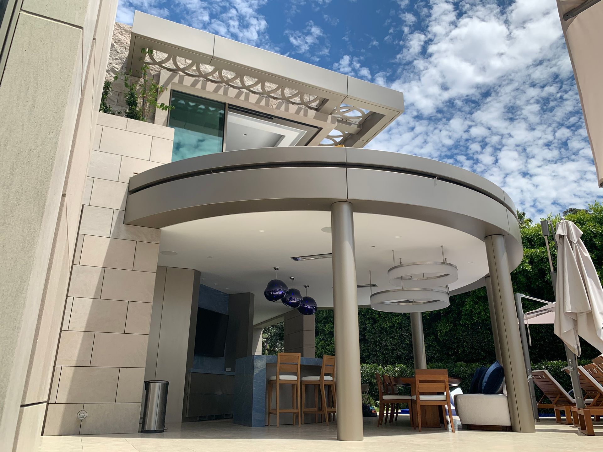 A modern outdoor patio with a curved roof, bar stools, dining set, and hanging blue lights under a blue, cloudy sky.