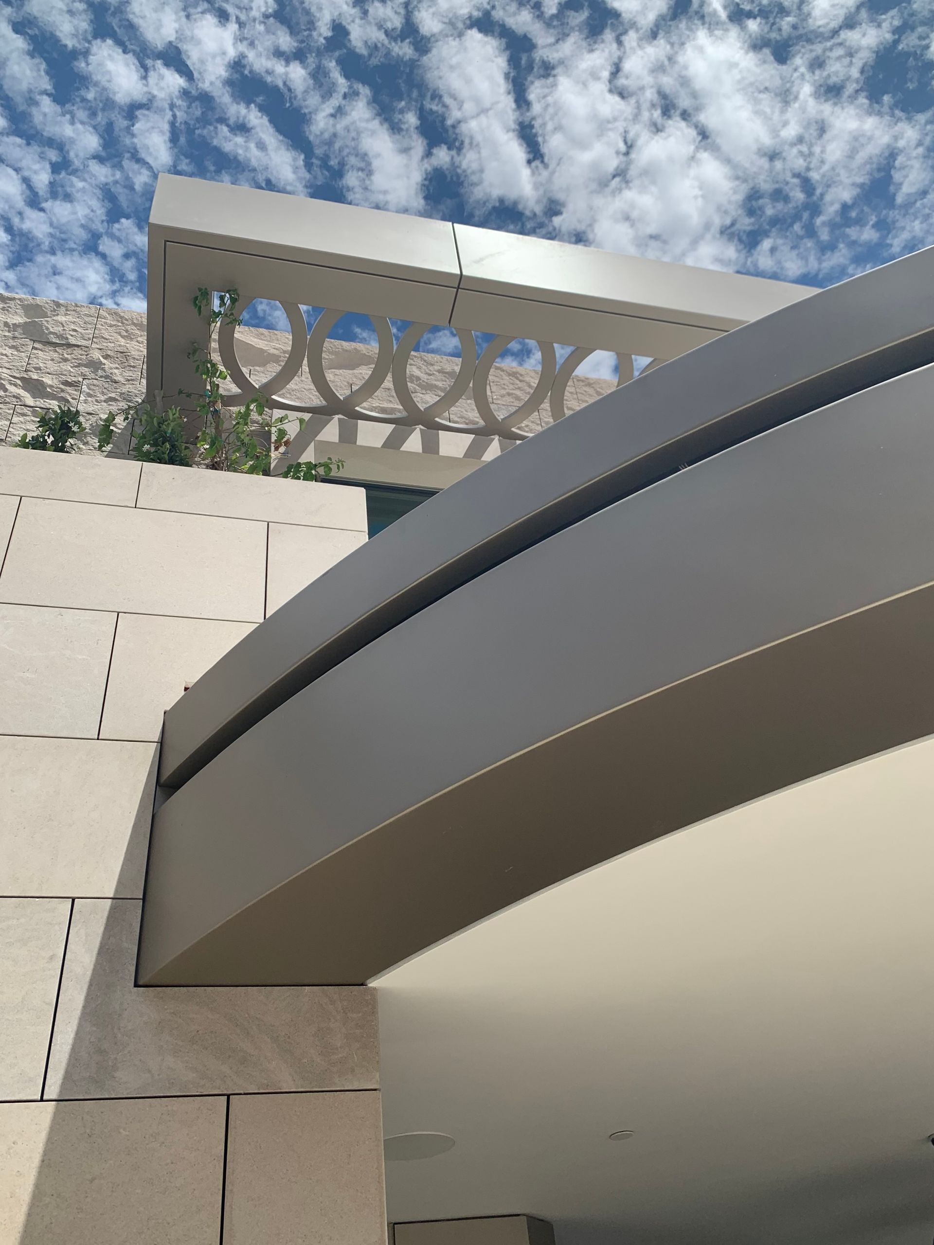 A low-angle view of a modern building with beige stone masonry, a dark metal overhang, and a decorative lattice screen.