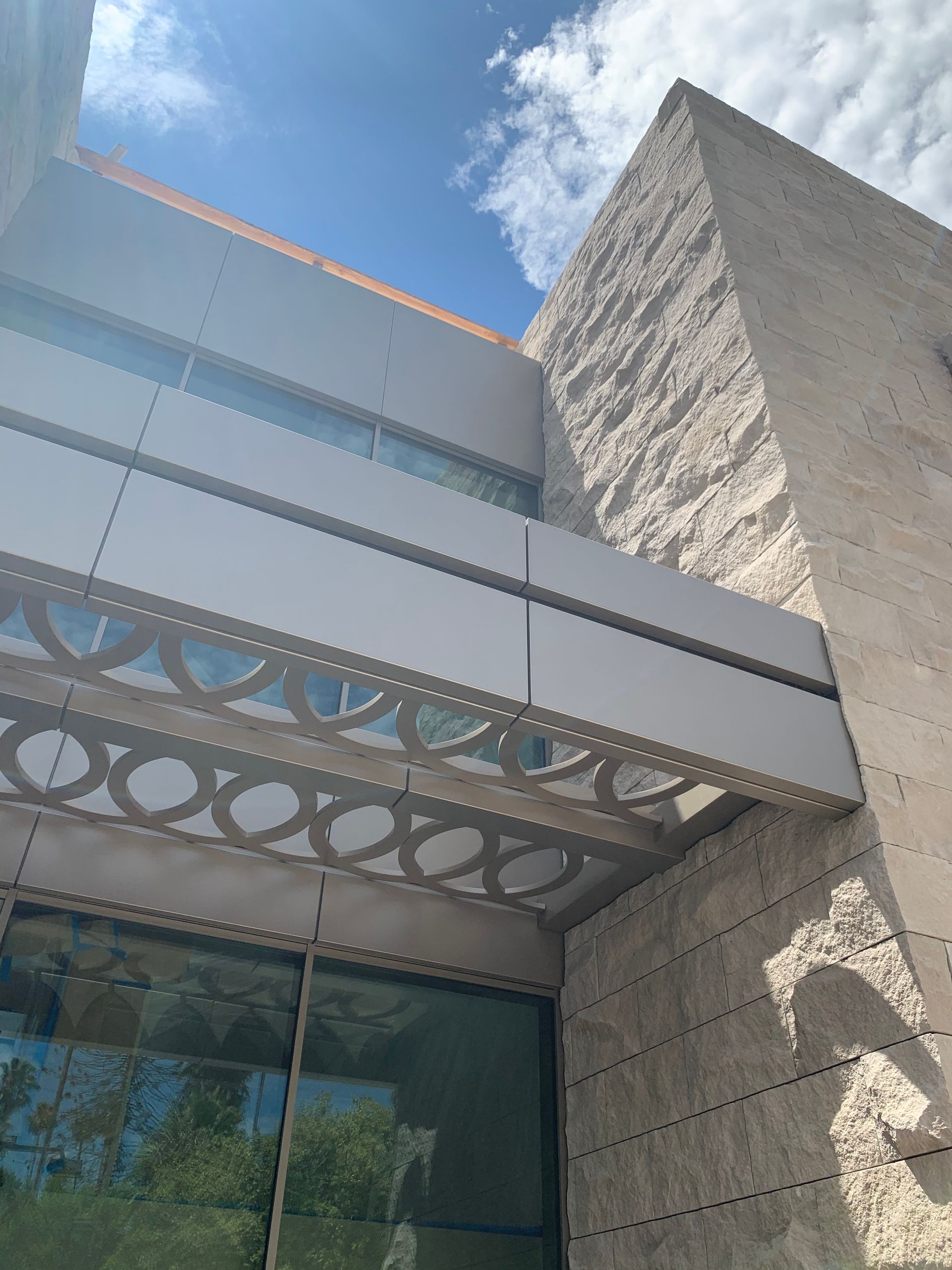 Modern building exterior with a textured stone column, geometric patterned overhang, and glass facade under a blue sky.