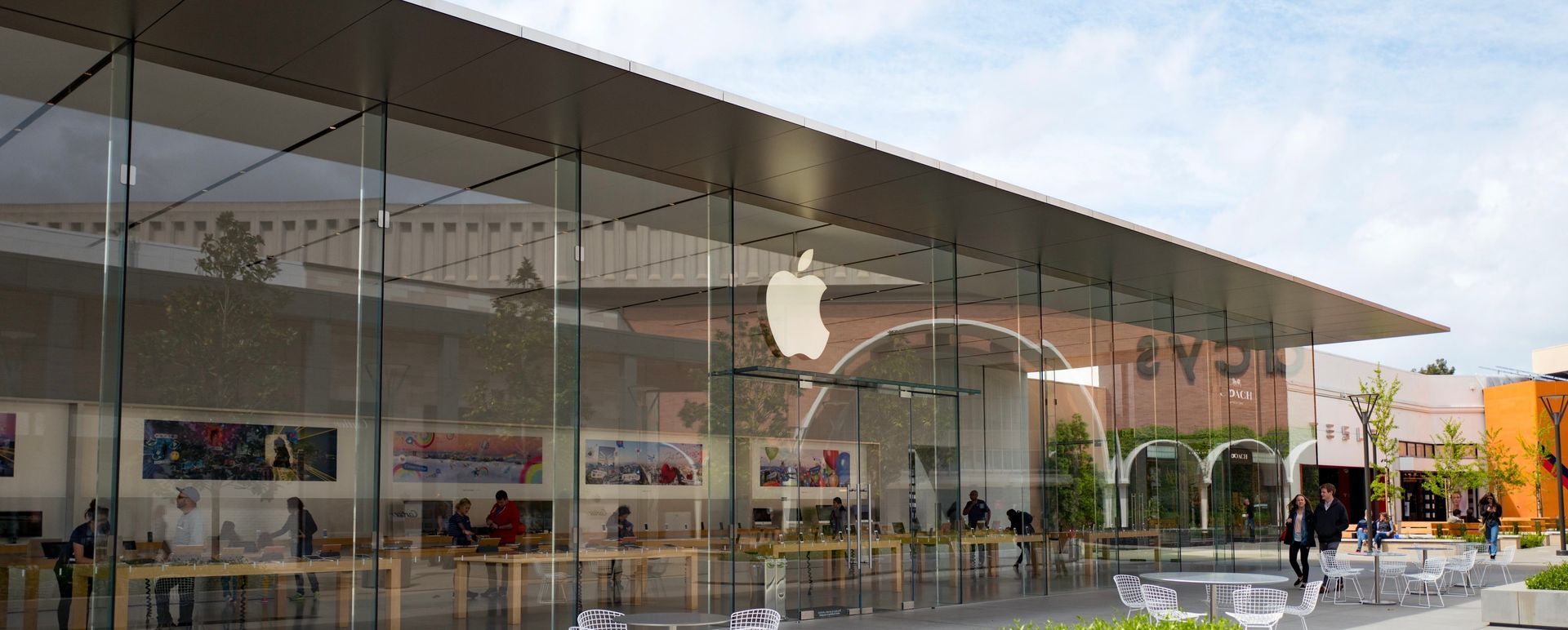 Modern Apple Store storefront with a glass facade, silver roof, and the iconic white Apple logo in an outdoor mall setting.