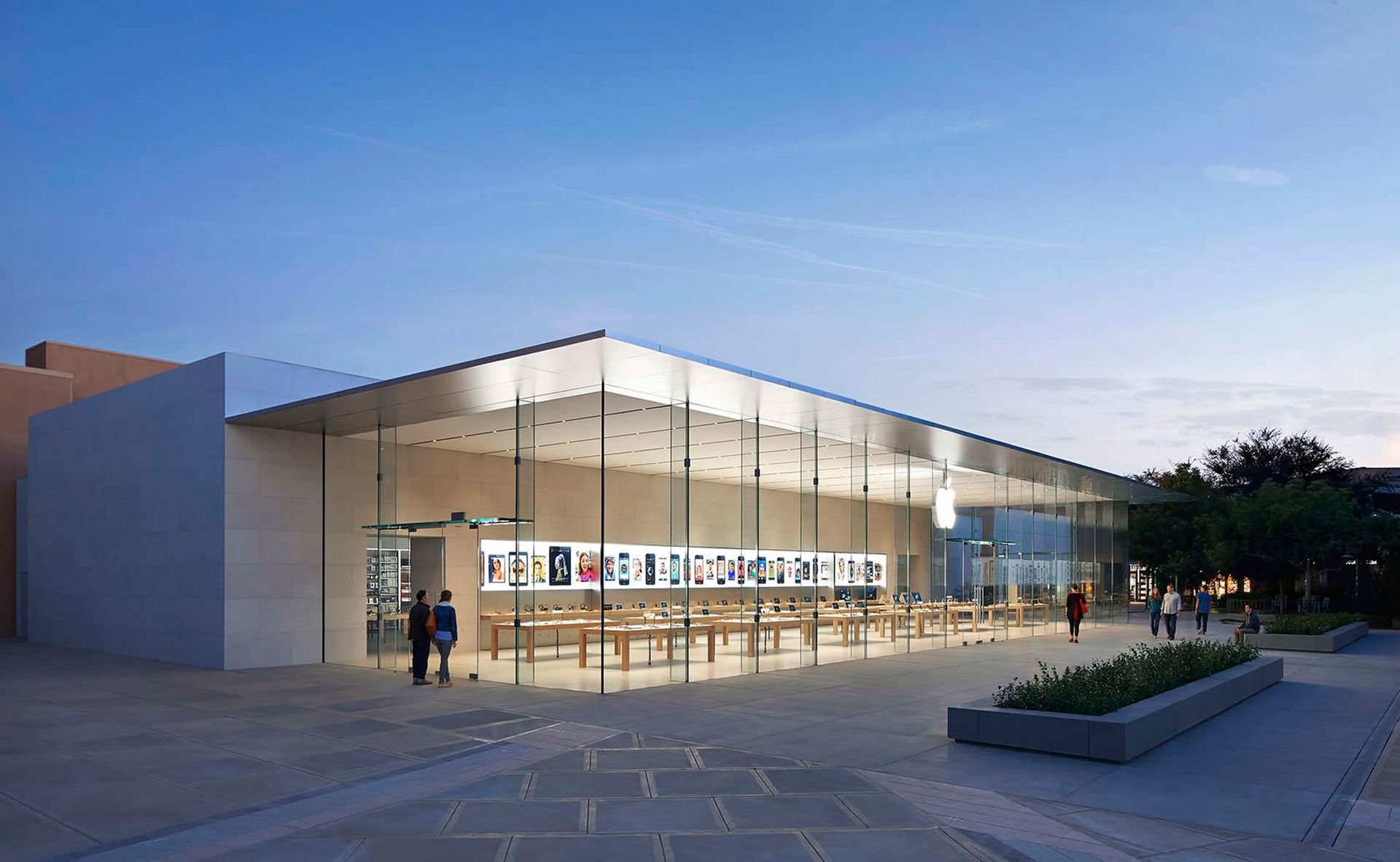 A wide, modern Apple Store with floor-to-ceiling glass walls, minimalist architecture, and a glowing logo at twilight.