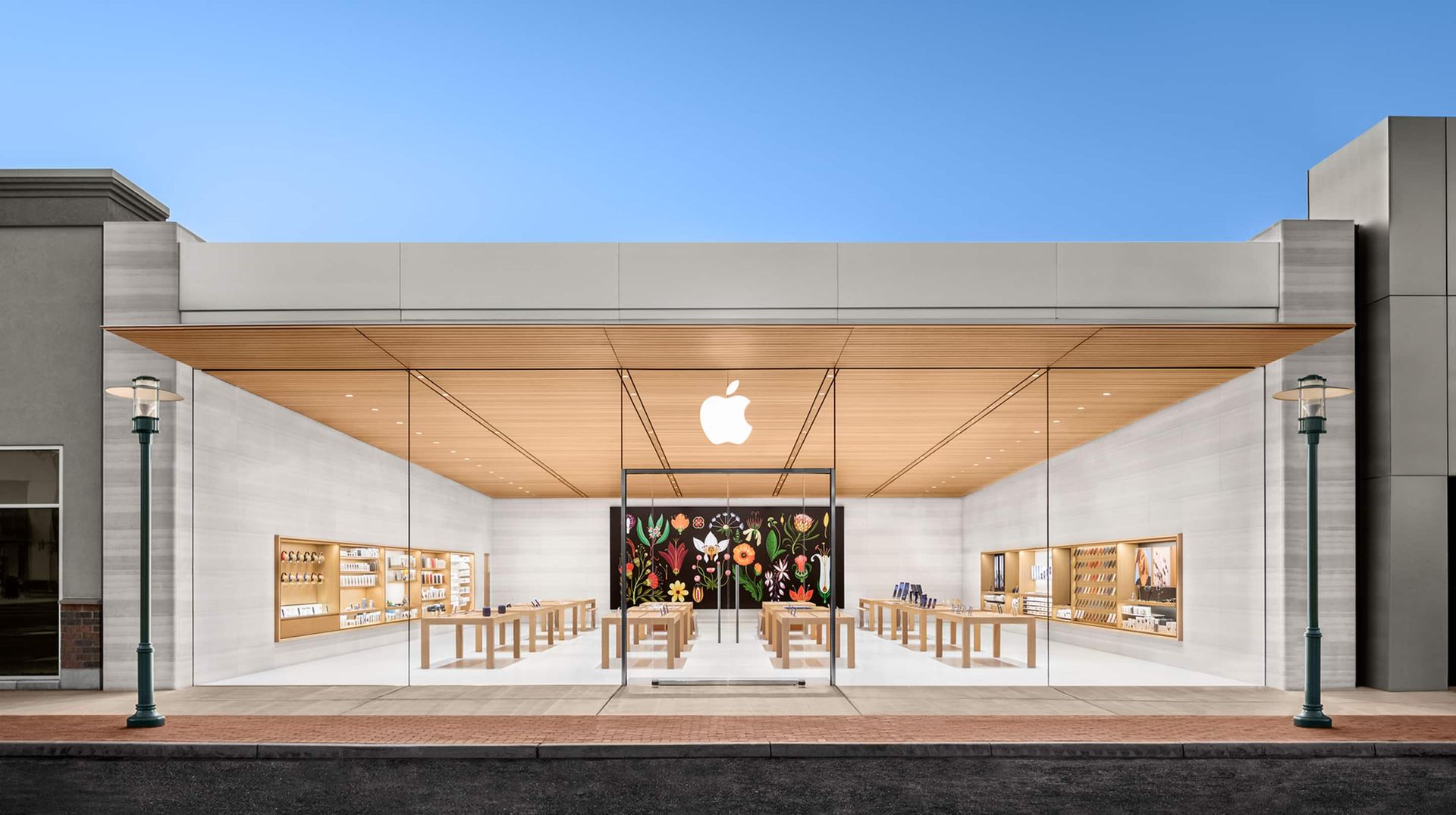 Exterior view of a bright, modern Apple Store with floor-to-ceiling glass, a wooden ceiling, and a large mural inside.