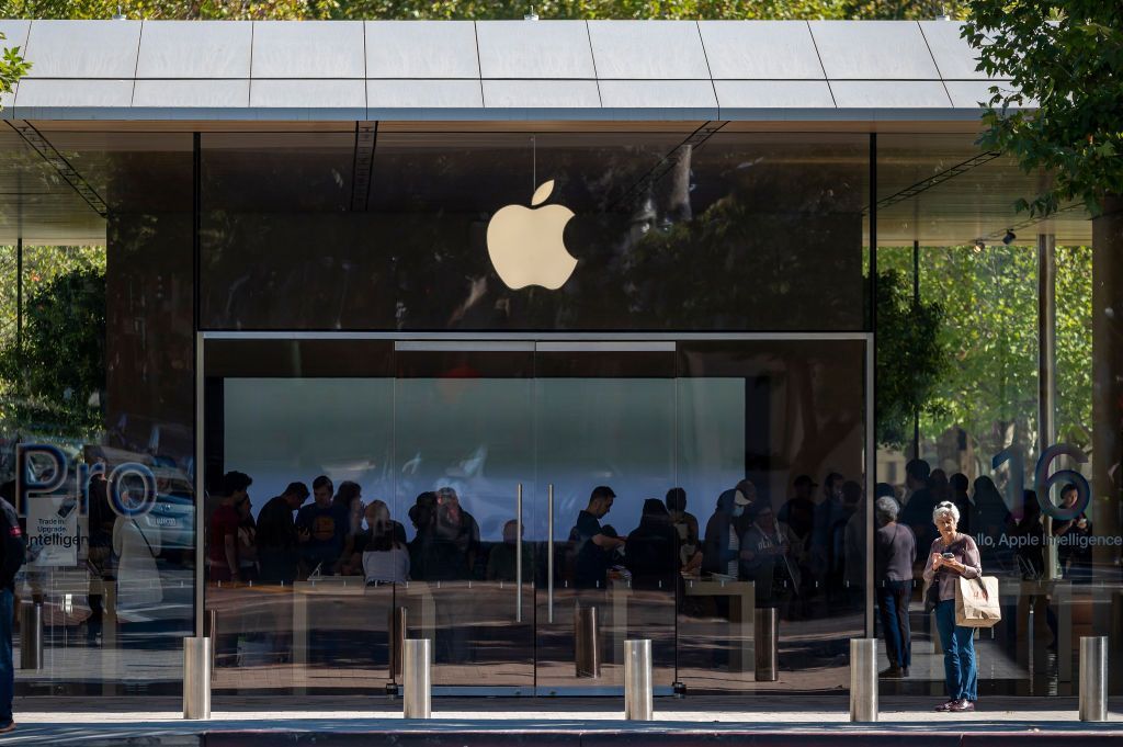 The glass-walled Apple store facade features a glowing logo above entrance doors with people inside and trees outside.