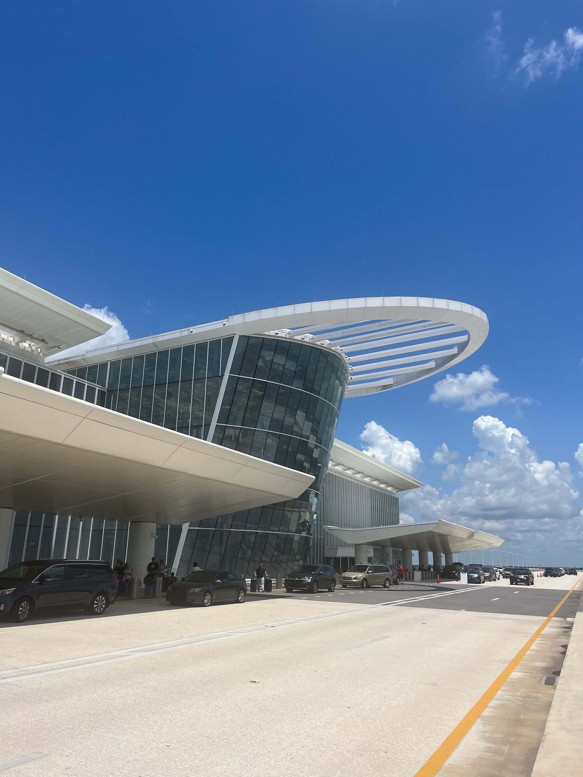 Modern airport terminal exterior with a curved glass facade, large white canopy, and cars parked in the drop-off area.