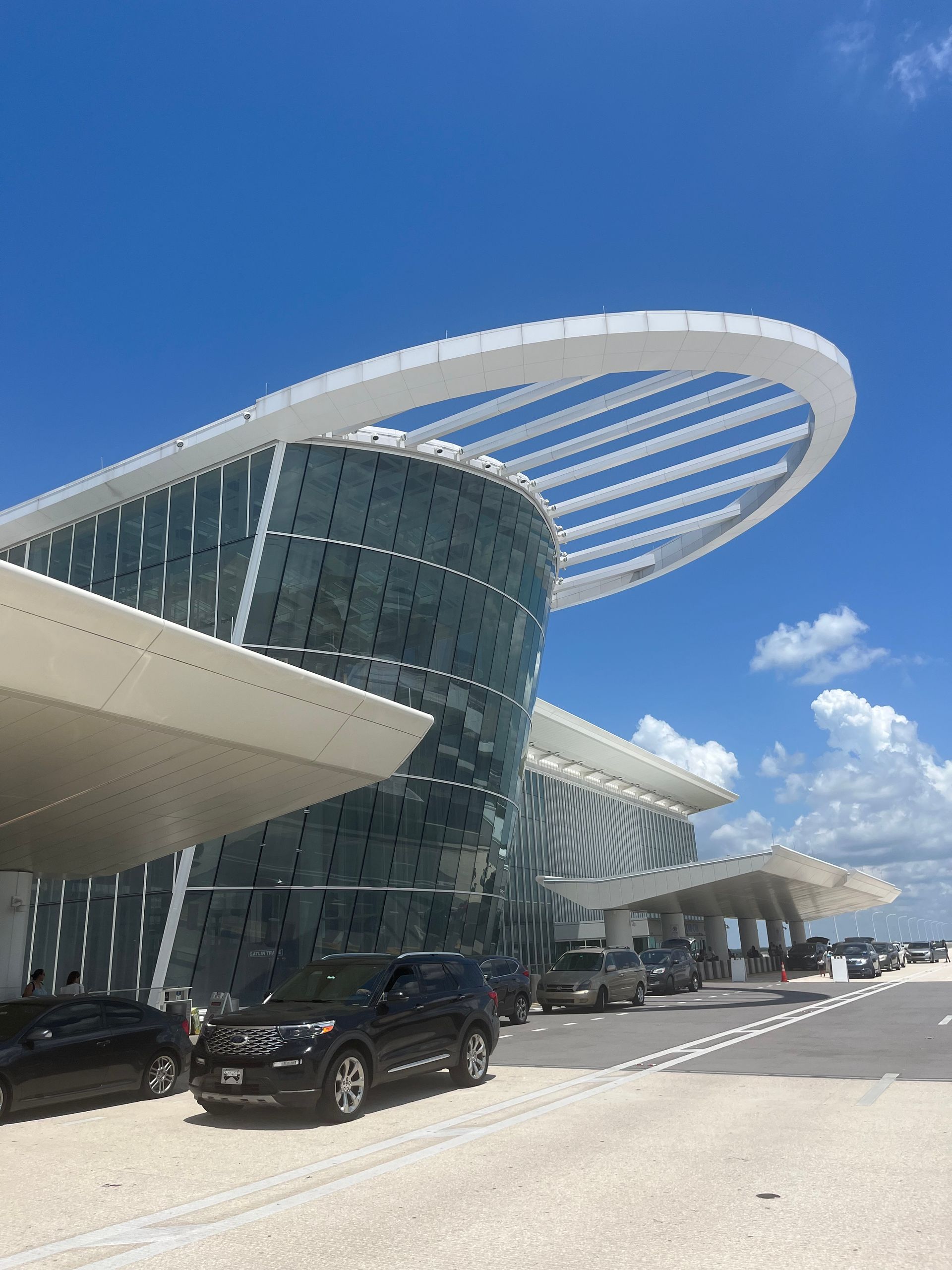 Modern glass terminal with curved white roof under a blue sky, with parked cars outside