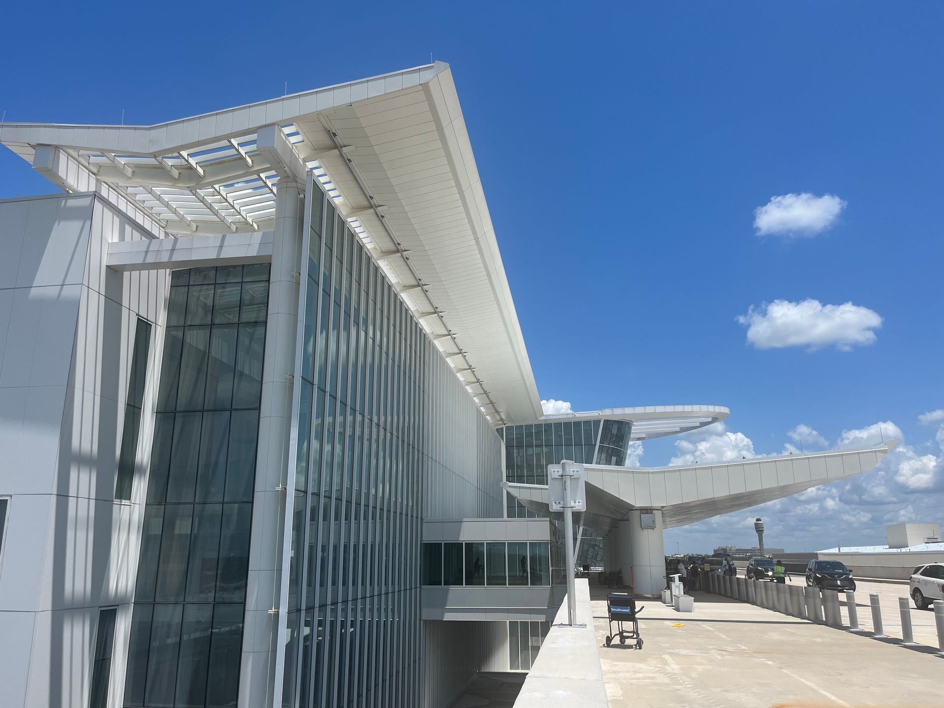 Modern airport terminal exterior with white structures, large glass windows, and overhanging roofs against a blue sky.