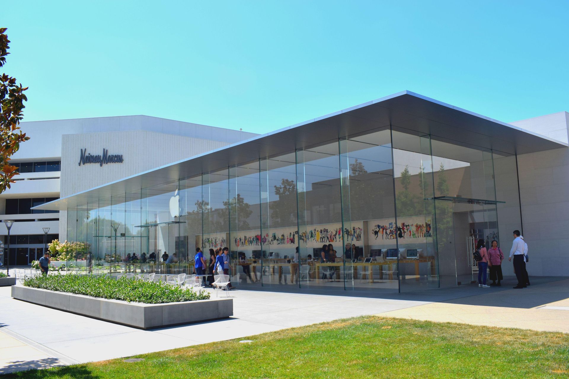 An Apple Store with a large glass facade next to a Neiman Marcus at an outdoor shopping center on a sunny day.