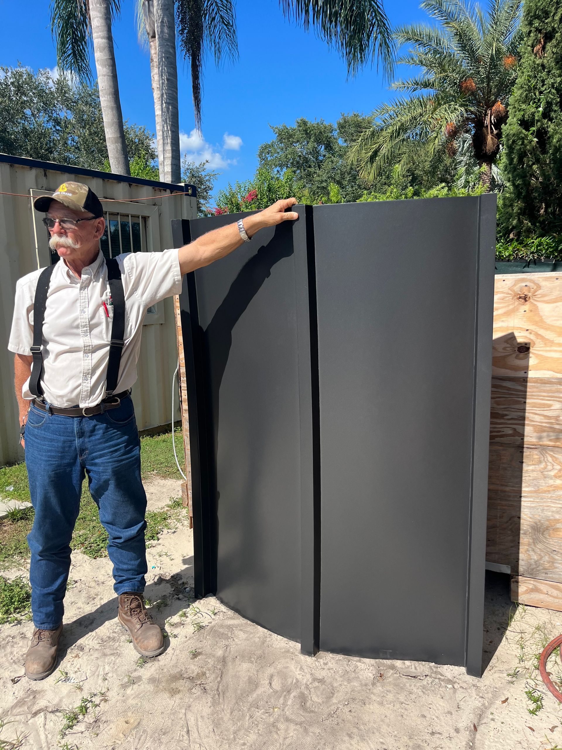 A person standing next to two tall, dark grey, vertical metal panels in an outdoor area with trees in the background.