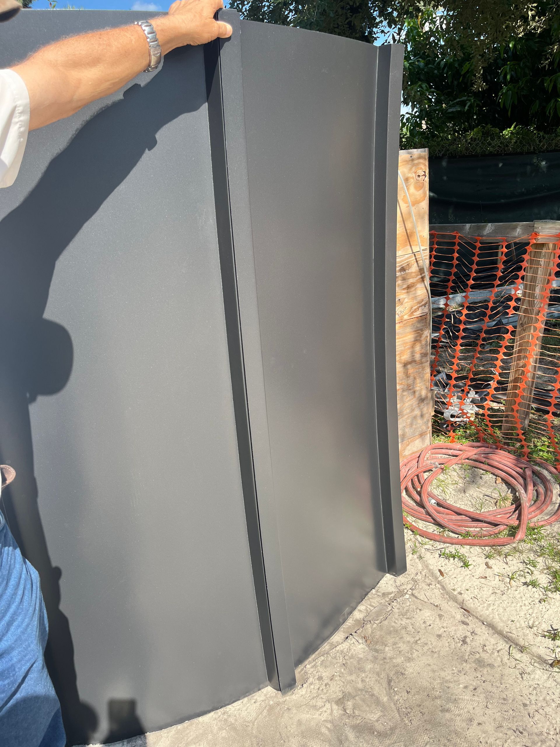 A person holds a large, matte-grey metal panel with a vertically notched edge, standing outdoors on a concrete surface.