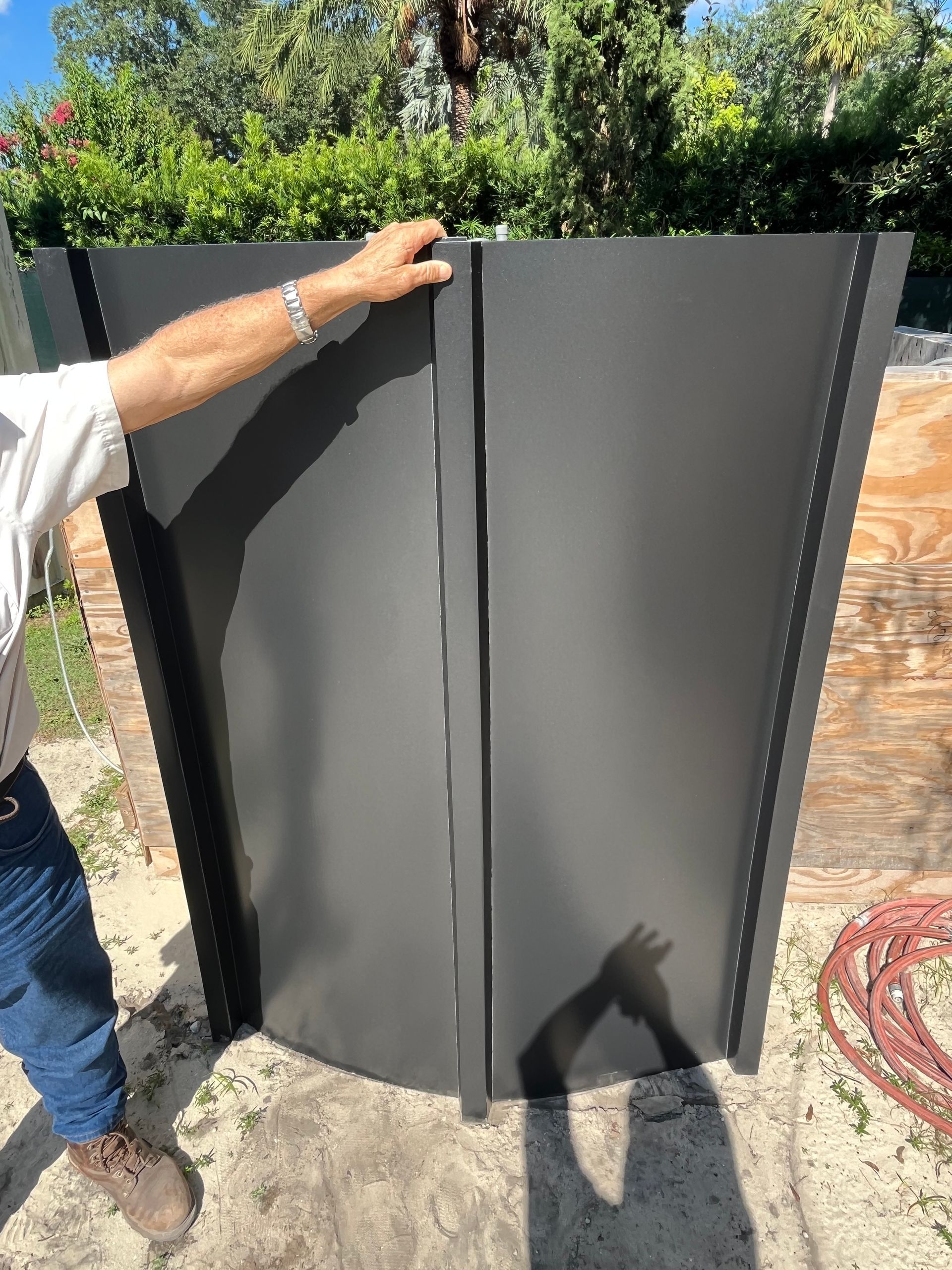 A person standing outdoors holds two matte-black, vertical metal panels together, showing their joint alignment.