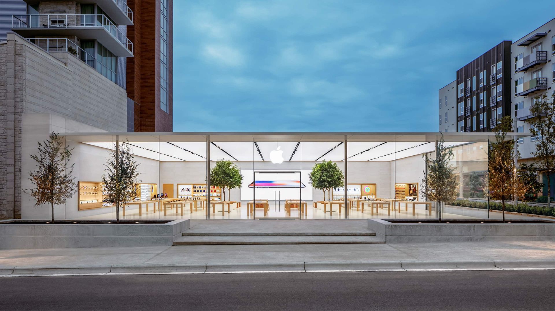 A modern, glass-walled Apple store at dusk with interior lighting illuminating wooden display tables and indoor trees.