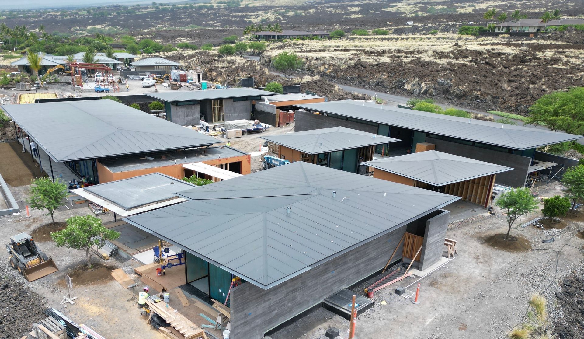 An aerial view shows multiple modern, dark-roofed buildings under construction in a dry, volcanic landscape.