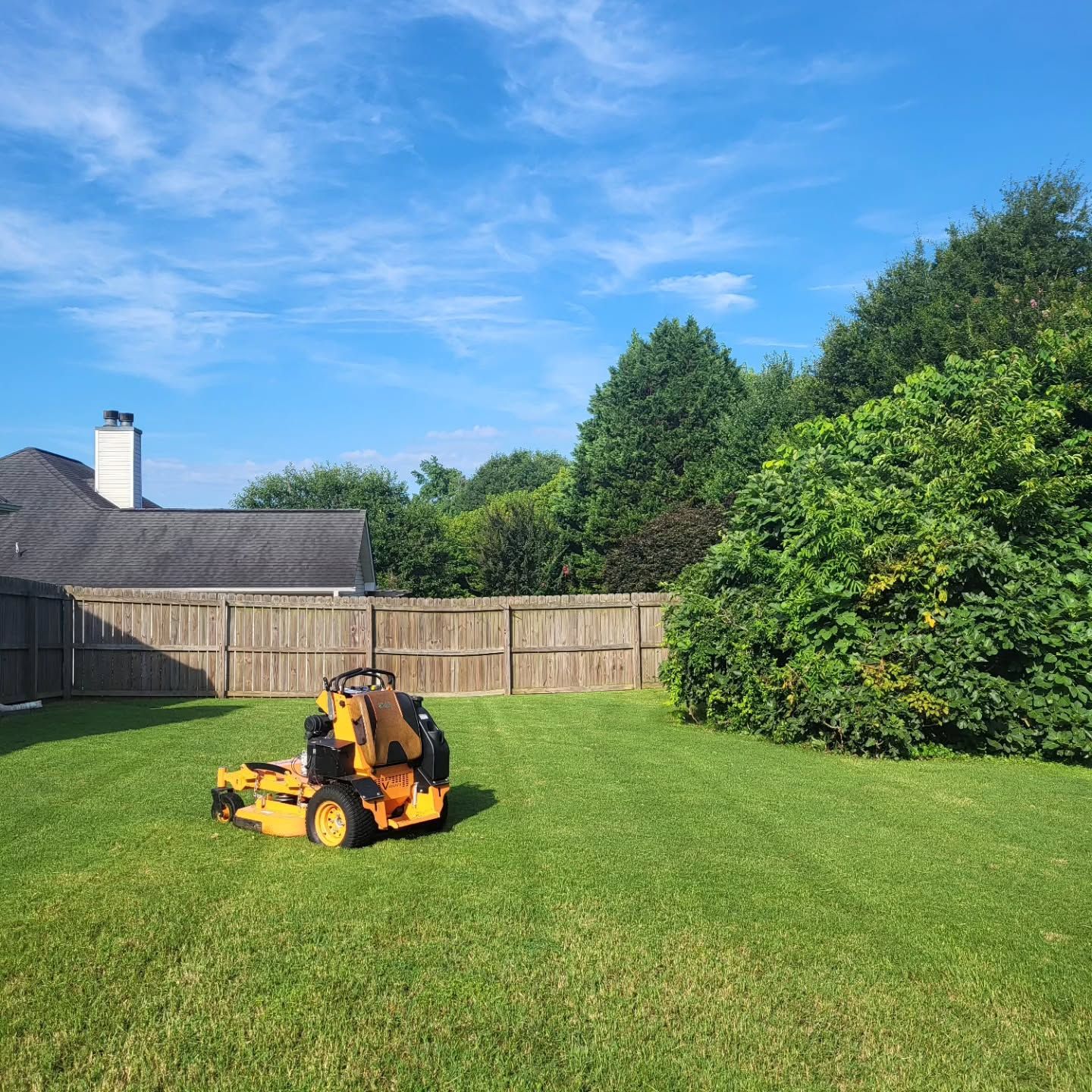 A yellow zero-turn lawn mower sits on a green residential lawn in front of a wooden fence and a house under a blue sky.