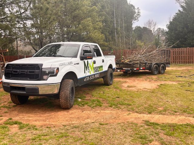 A white Ford pickup truck with branded decals is parked on grass, towing a trailer filled with tree branches.