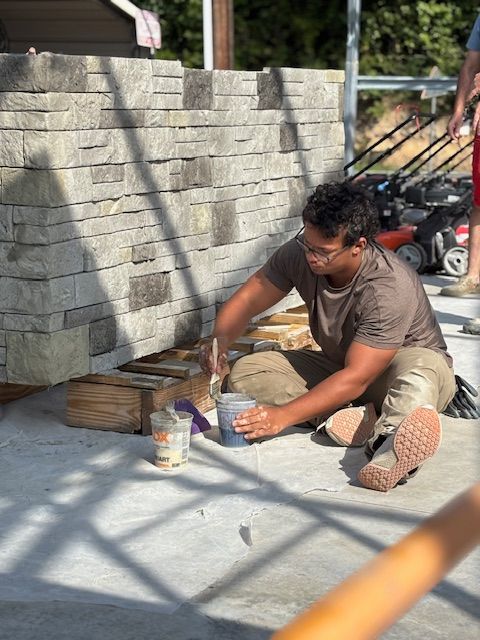 A person sits on a concrete floor, applying sealant with a brush to the base of a stone veneer wall.
