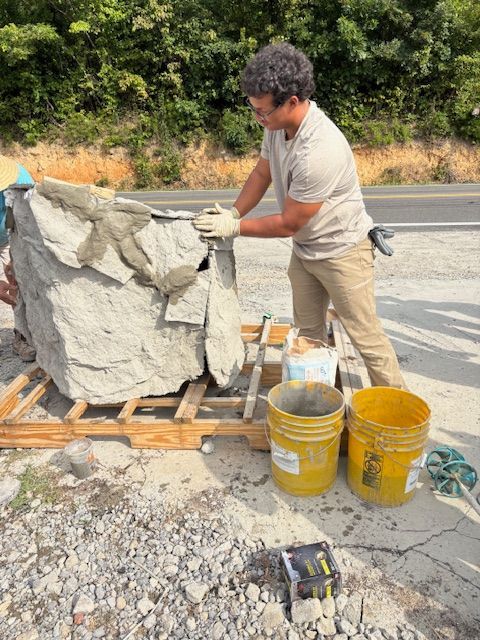 A person wearing work gloves applies mortar to a large rock on a wooden pallet next to two yellow buckets.