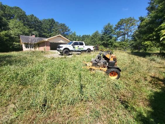 A commercial mower clears a large grassy lot with a white work truck and a house in the background.