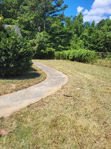 A curved concrete sidewalk cuts through a dry, grassy lawn toward a line of green trees under a blue, cloudy sky.