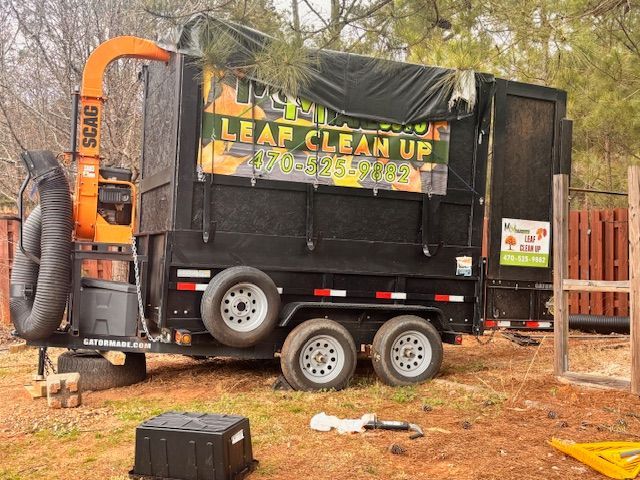 A black leaf removal trailer with a mounted orange Scag leaf vacuum, parked on a dirt lot near a wooden fence.