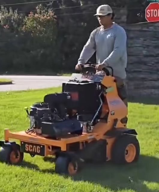 A person stands behind a bright orange Scag stand-on commercial lawn mower on a grassy lawn near a stop sign.