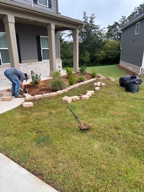 A person in a gray shirt kneels to place stone landscape edging along a flower bed in front of a house.