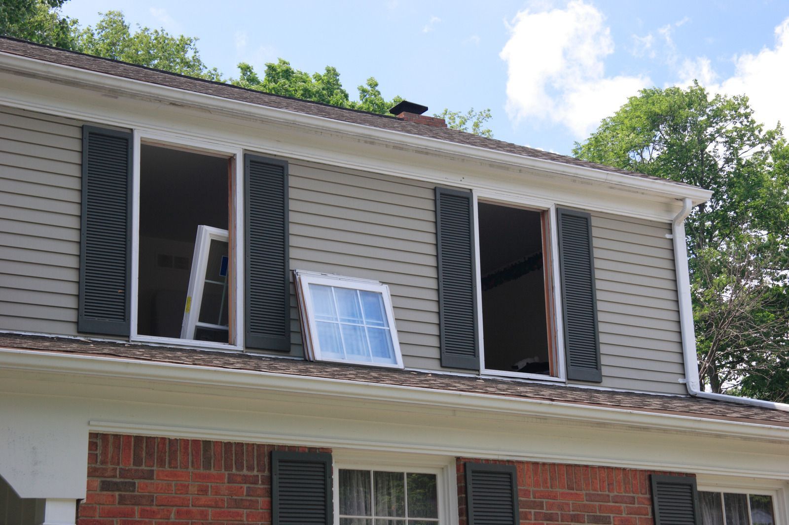 House exterior with open windows, gray siding, black shutters, and brick base. A new window is propped outside.