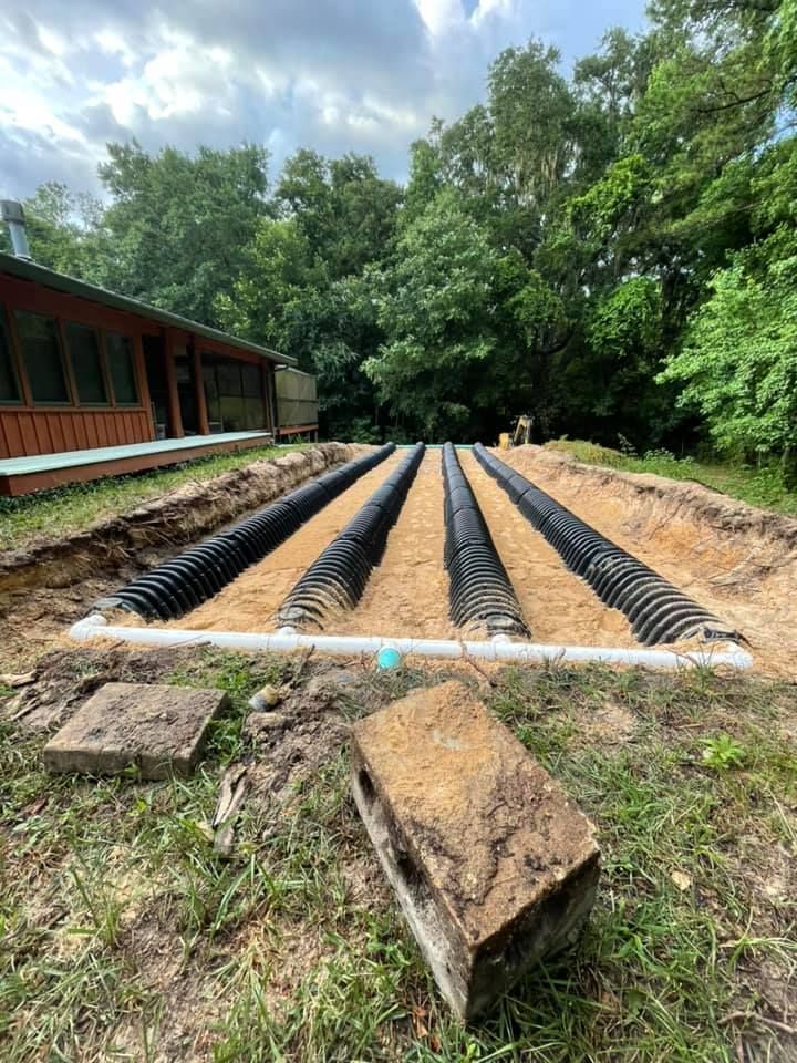 Septic system installation: black pipes laid in trenches filled with sand next to a house and trees.