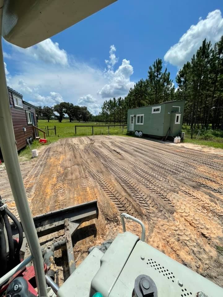 A green tiny house trailer on a cleared sandy lot under a blue sky, with other small structures visible.