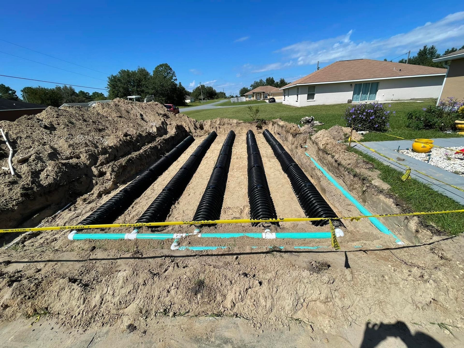 Septic system installation in progress: black drain lines in trenches with tan sand, green pipes, dirt, and houses in the background.