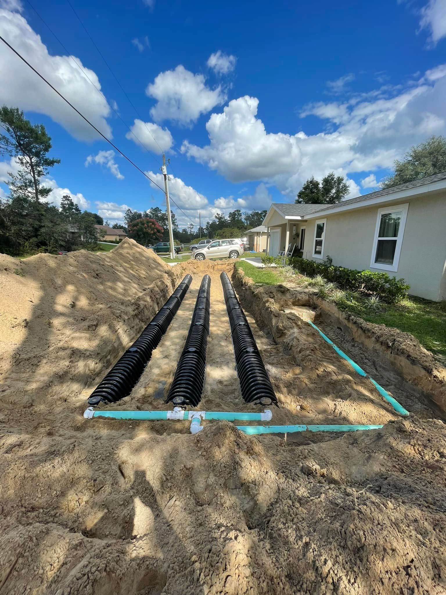 Rows of black septic tanks installed in a sandy trench under a blue sky, near a house.