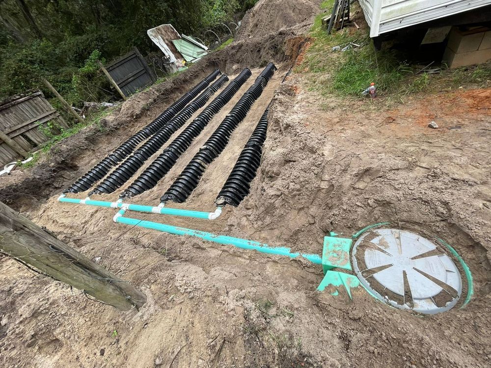 Septic system installation; black corrugated pipes, turquoise pipes, and a round access lid in a dirt trench.