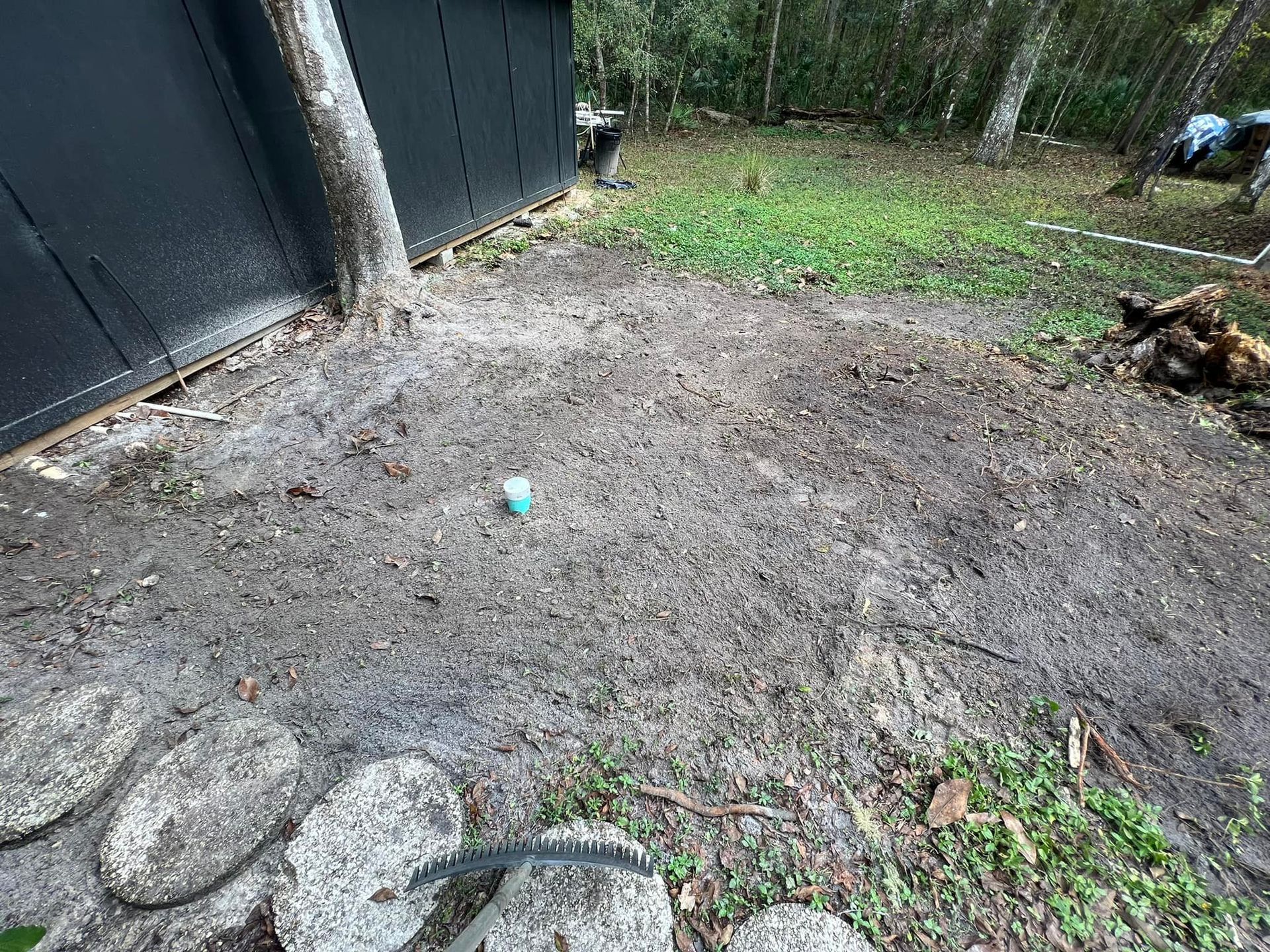 A dirt patch next to a black structure, with stepping stones in the foreground and greenery in the distance.