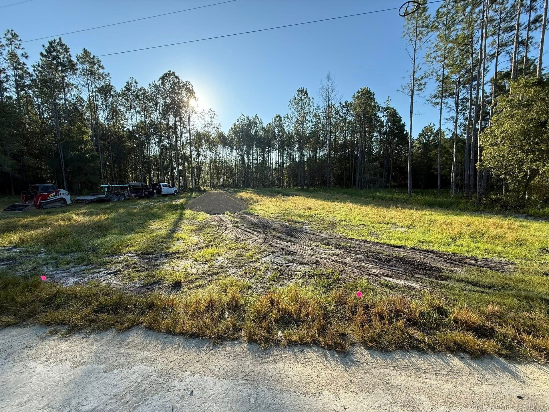 Cleared lot with dirt pile, grass, trees, and construction equipment under a sunny sky.