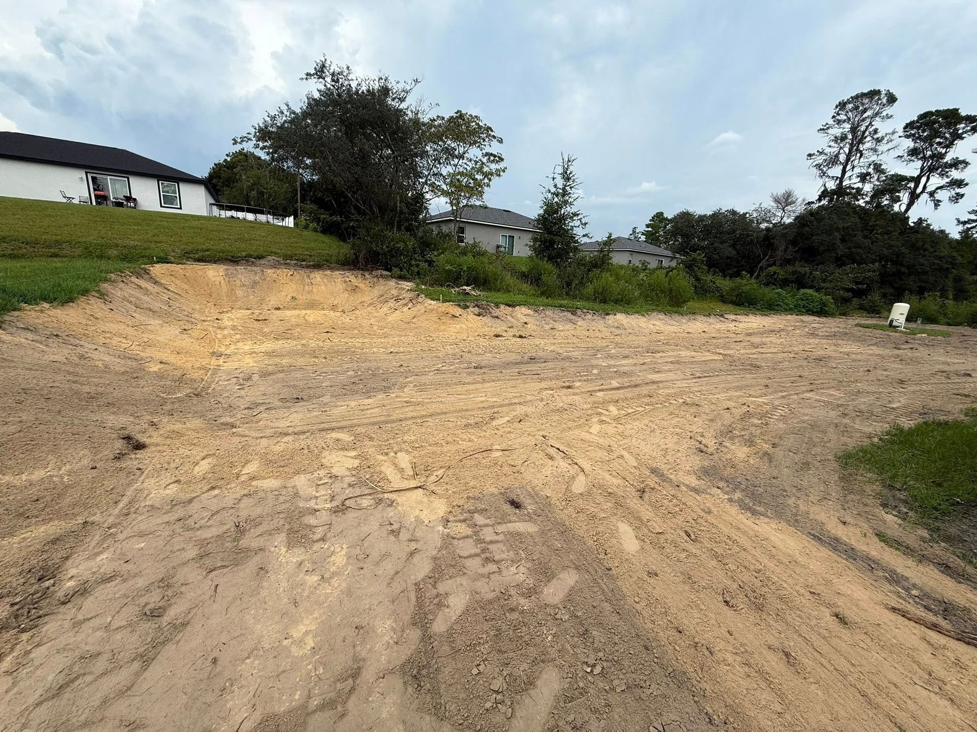 Dirt lot with tire tracks and some vegetation. Houses and trees in the background under cloudy sky.